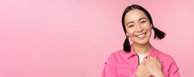Carina ragazza coreana con capelli sciocchi che sembrano felici e grati grazie posa tenendosi per mano sul cuore lusingato in posa su sfondo rosa