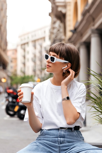 Carina ragazza dai capelli scuri in occhiali da sole che beve caffè in una buona giornata di sole Ritratto di modello femminile jocund in abito elegante che tiene il caffè