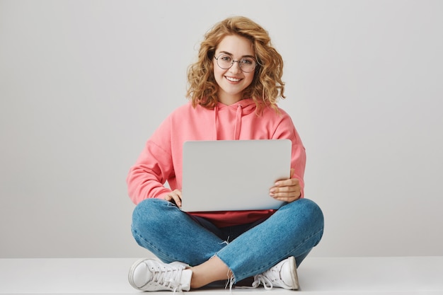 Carina ragazza freelance utilizzando laptop, seduta sul pavimento e sorridente