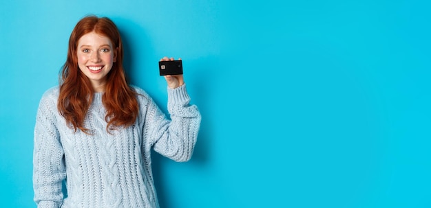 Carina ragazza rossa in maglione che mostra la carta di credito sorridendo alla telecamera in piedi su sfondo blu