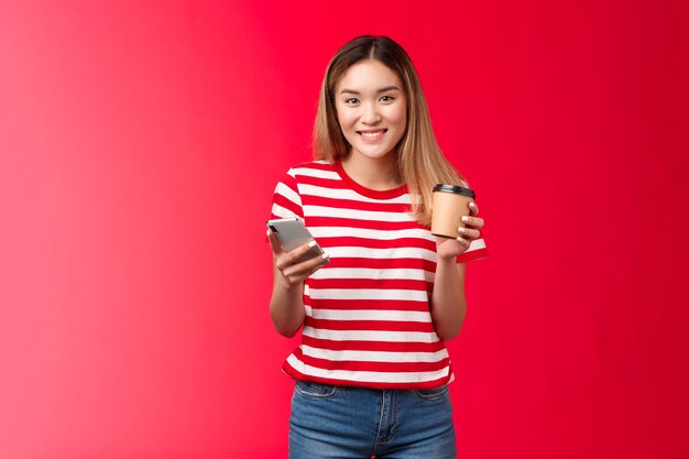 Carina studentessa universitaria asiatica bionda in piedi in pausa pranzo godersi un caffè vicino al campus tenere lo smartphone ...