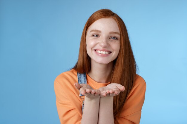 Carina tenera gentile giovane ragazza allo zenzero che dà a tutti ti amo, tieni qualcosa sui palmi che mostrano la macchina fotografica sorridente felice di presentare il presente ghignante gesto romantico invia baci d'aria, sfondo blu