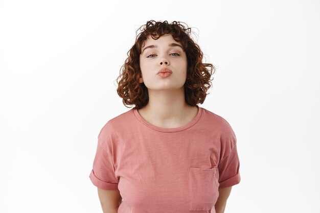 Carino bellissimo modello femminile che si appoggia sulle labbra per il bacio, vuole baciare, sembra romantico e sciocco, in piedi in t-shirt su sfondo bianco. Concetto di relazione e romanticismo