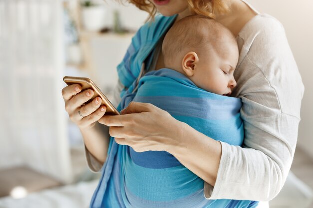 Carino piccolo bambino che fa un sonnellino pacificamente mentre la madre lo abbraccia e manda un sms al marito per telefono chiedendo di comprare un po 'di alimenti per bambini e pannolini. Stile di vita, concetto di famiglia.