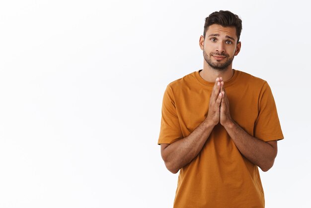 Carino sciocco attraente hipster uomo in maglietta marrone che fa promessa sorridente con angelico sorriso innocente premere le mani insieme sul petto pregando o chiedendo aiuto hanno bisogno di qualcosa di sfondo bianco