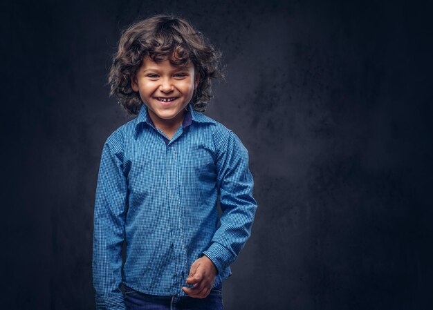 Carino scolaro sorridente con capelli ricci castani vestito con una camicia blu, in posa in uno studio. Isolato su uno sfondo scuro con texture.