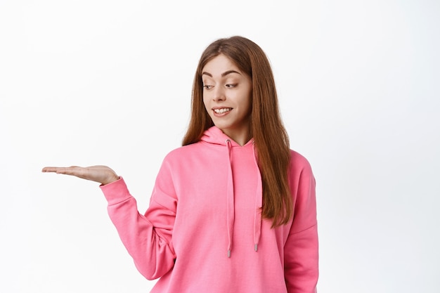 Carino studentessa adolescente, tenendo copyspace sul palmo, mostra il prodotto in mano contro il muro bianco, sorridendo e guardando l'oggetto, muro bianco