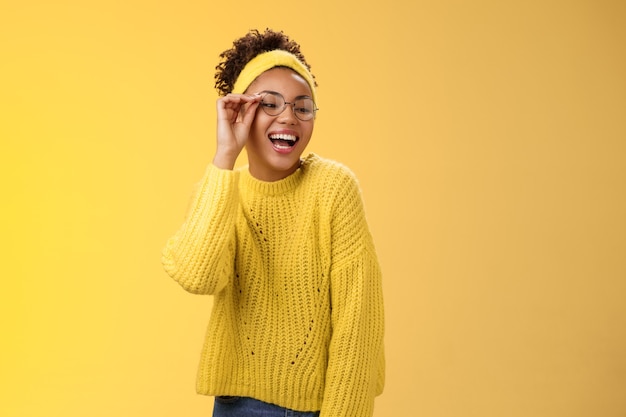 Carismatico elegante fiducioso bella ragazza adolescente gioiosa afro-americana che tocca gli occhiali naso che ride ampiamente guarda in basso nell'angolo destro sorridendo divertiti scherzando godendo, sfondo giallo.