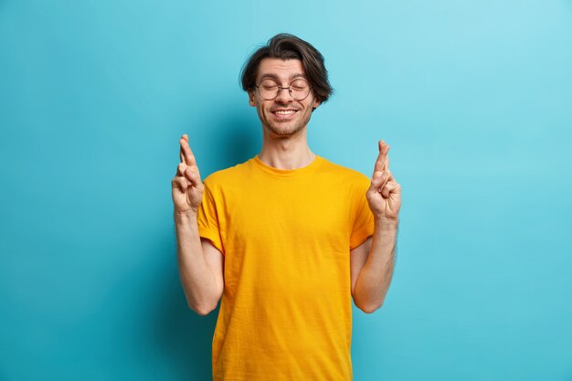 Caro concetto di desiderio. Il ragazzo positivo con l'acconciatura alla moda incrocia le dita in pose con gli occhi chiusi prega per miracoli sperando che accadano cose buone