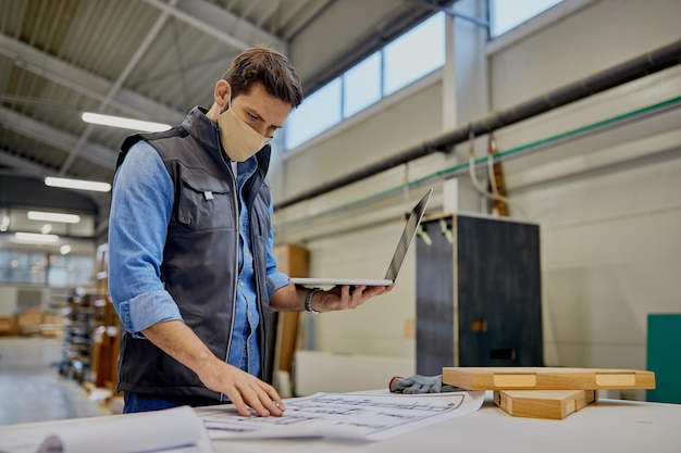 Carpentiere con maschera facciale che esamina i progetti durante l'utilizzo del computer presso l'impianto di produzione di lavorazione del legno