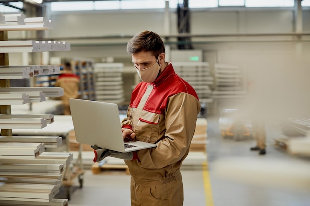 Carpentiere maschio con maschera facciale che usa il laptop mentre controlla le scorte in un'officina