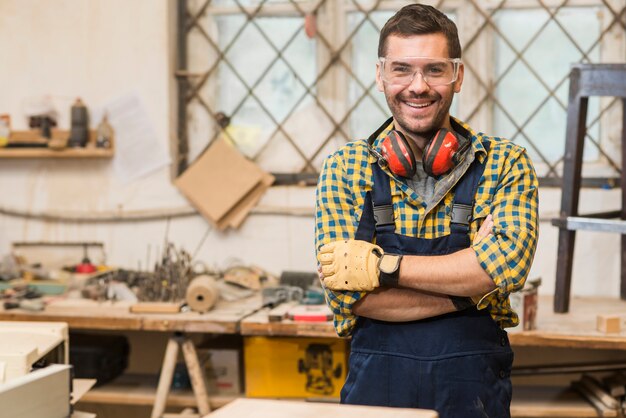 Carpentiere maschio sorridente che indossa gli occhiali di protezione che stanno davanti al banco da lavoro con il suo braccio attraversato