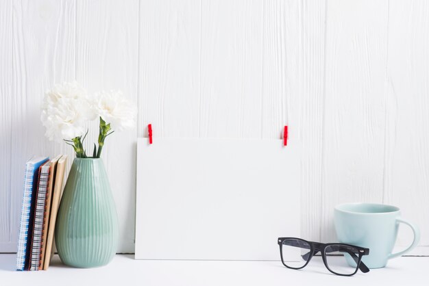 Carta bianca bianca con molletta rossa; occhiali; tazza; vaso e libri sul contesto strutturato in legno
