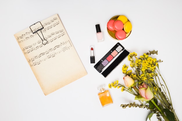 Carta per appunti musicale d&#39;epoca; rossetto; bottiglia di smalto per unghie; bottiglia di profumo; bouquet di fiori e amaretti su sfondo bianco