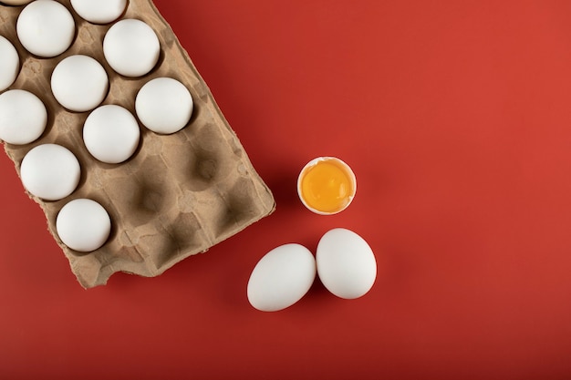 Cartone di uova bianche con tuorlo su superficie rossa.