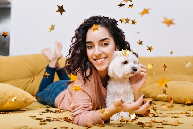 Casa momenti felici con animali domestici di giovane bella donna con capelli ricci castana tagliati divertendosi in orpelli dorati sul divano in appartamento moderno. Modello grazioso adorabile che si rilassa a casa con il piccolo cane bianco