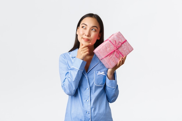 Casa, vacanze e concetto di stile di vita. Intrigante ragazza di buon compleanno in pigiama blu che agita la scatola con regalo per scoprire cosa c'è dentro, indovinando il presente e sorridendo curioso, sfondo bianco