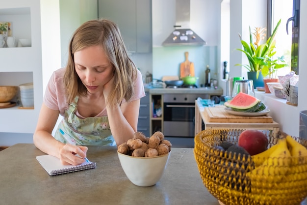 Casalinga concentrata che pianifica il menu settimanale nella sua cucina, annotando la lista della spesa nel taccuino. Cucinare a casa concetto