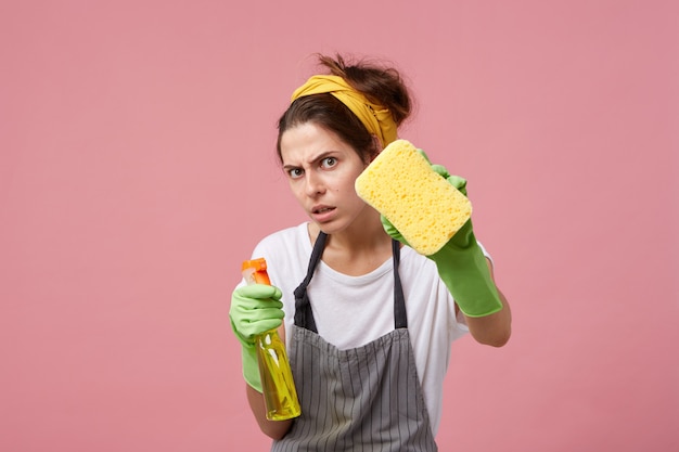 Casalinga disperata e scrupolosamente ordinata lava i vetri con spugna e detergente spray mentre fa le pulizie in casa. Giovane donna europea che indossa guanti di gomma verde che riordina durante il fine settimana