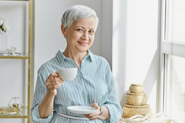 Casalinga matura dai capelli grigi elegante attraente che porta vestito blu alla moda, in piedi vicino alla finestra con una tazza di caffè mentre pranza o fa colazione. Persone, stile di vita e concetto di ospitalità