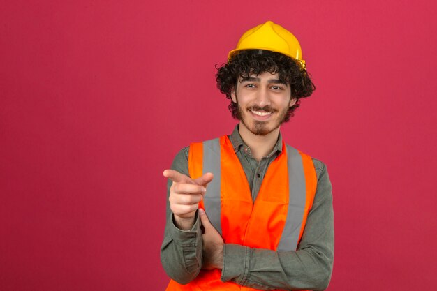 Casco e giubbotto di sicurezza d'uso del giovane ingegnere bello barbuto che sorridono allegramente indicando qualcosa con il dito sopra la parete rosa isolata