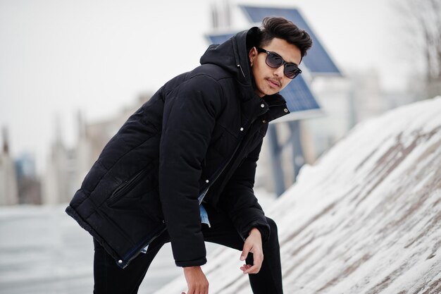 Casual giovane indiano in giacca nera e occhiali da sole poste al giorno d'inverno