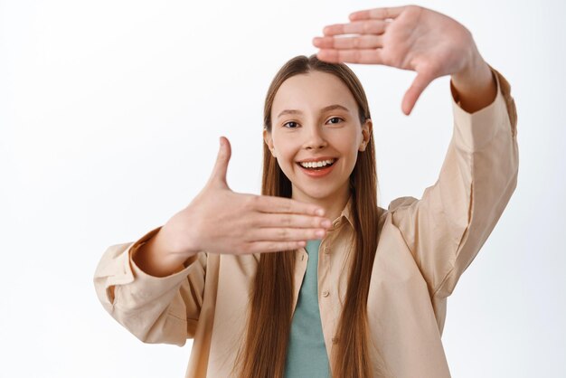 Cattura il momento La ragazza sorridente che fa le mani incornicia il gesto della fotocamera e guarda attraverso la ricerca di un angolo perfetto per l'immagine di qualcosa in piedi su sfondo bianco