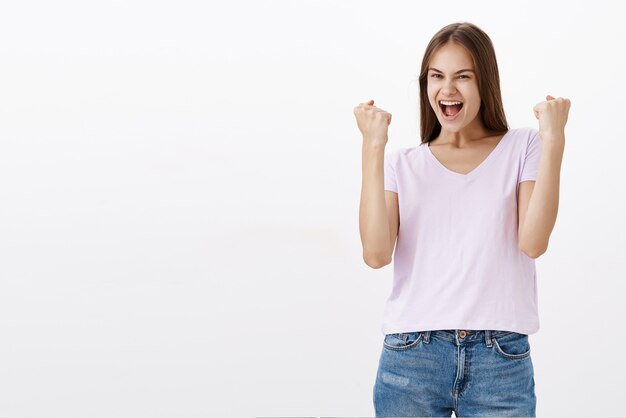 Celebrando e trionfando felice ragazza di bell'aspetto che alza i pugni chiusi in gesto di sì gioendo per il raggiungimento di un buon risultato sentendosi gioioso dalla vittoria sul muro bianco