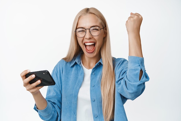 Celebrando la donna fortunata sveglia che vince sul telefono cellulare che tiene smartphone e trionfa tifo e sorridente in piedi su sfondo bianco