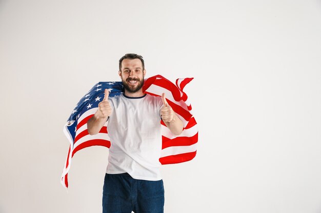 Celebrare un giorno dell'indipendenza. Stelle e strisce. Giovane con la bandiera degli Stati Uniti d'America isolato sulla parete bianca dello studio. Sembra follemente felice e orgoglioso come un patriota del suo paese.