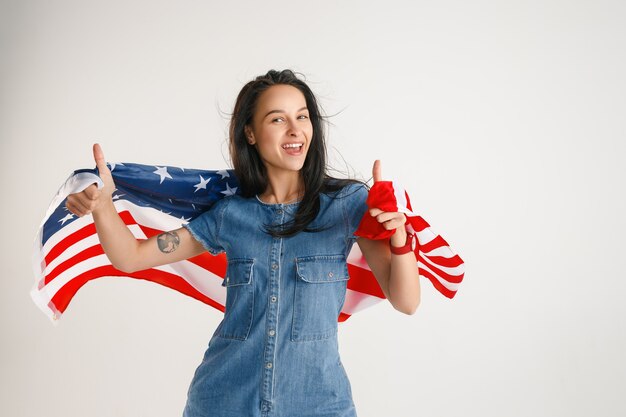 Celebrare un giorno dell'indipendenza. Stelle e strisce. Giovane donna con la bandiera degli Stati Uniti d'America isolato sulla parete bianca dello studio. Sembra follemente felice e orgogliosa come una patriota del suo paese.