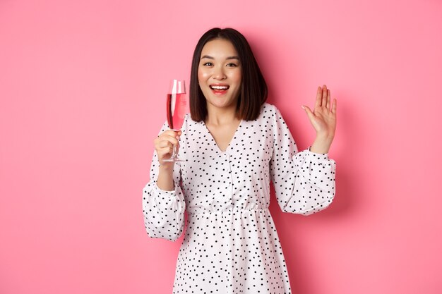 Celebrazione. Bella donna asiatica che beve champagne e sorride, in piedi in abito su sfondo rosa.