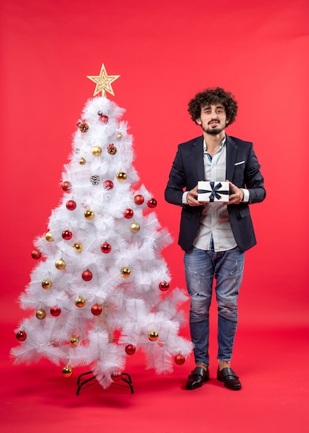 Celebrazione del nuovo anno con giovane serio che tiene un regalo vicino all'albero di Natale bianco decorato su metraggio rosso