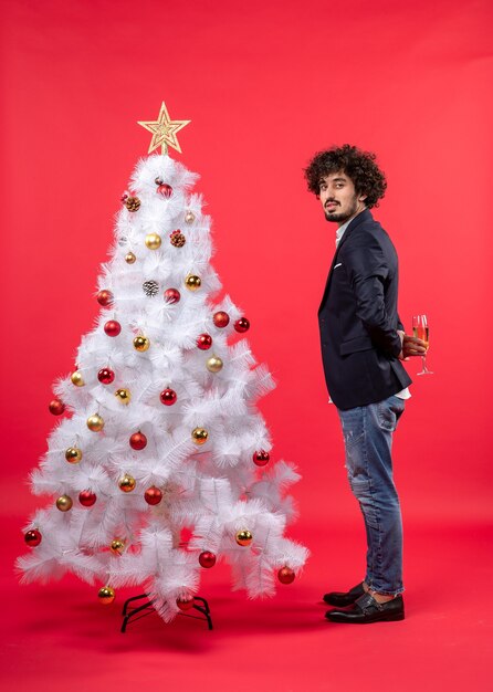 Celebrazione del nuovo anno con il giovane che tiene un bicchiere di vino dietro l'albero di Natale bianco decorato sul rosso