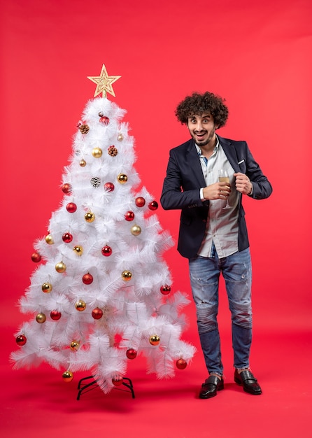 Celebrazione del nuovo anno con il giovane che tiene un bicchiere di vino vicino al suo cuore vicino all'albero di Natale bianco decorato