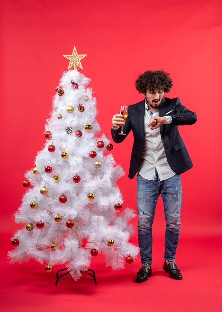 Celebrazione di Natale con il giovane barbuto con vino che controlla il tempo e in piedi vicino all'albero di Natale sul rosso