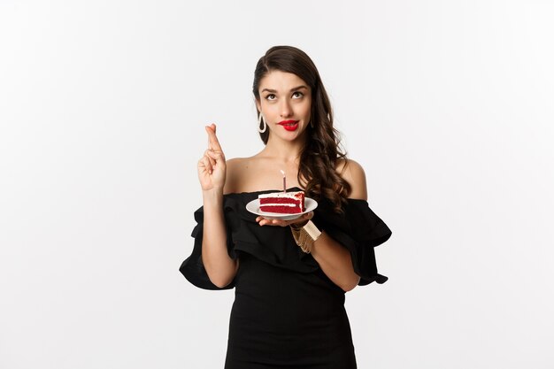 Celebrazione e concetto di festa. Donna speranzosa e sognante che esprime desiderio sulla torta di compleanno, incrociare le dita e sorridere felice, in piedi su sfondo bianco.