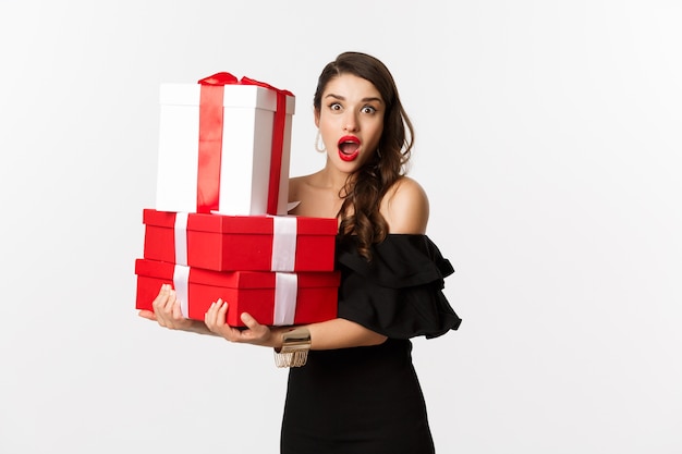 Celebrazione e concetto di vacanze di Natale. Bella donna in abito nero, tenendo doni e guardando sorpreso, in piedi su sfondo bianco.