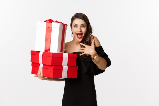 Celebrazione e concetto di vacanze di Natale. Una donna eccitata e felice riceve regali, tiene in mano regali di Natale e si rallegra, in piedi in abito nero su sfondo bianco
