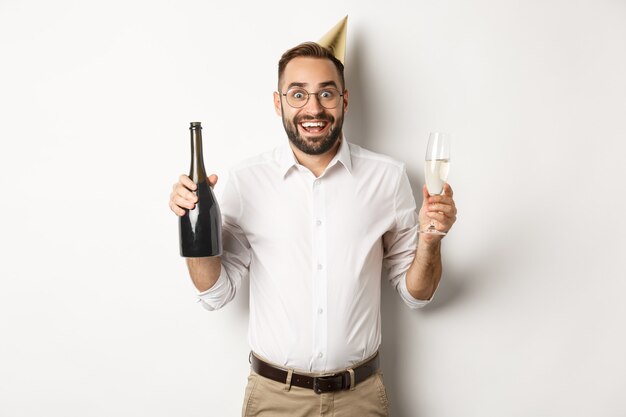 Celebrazione e festività. Uomo emozionante che gode della festa di compleanno, indossa il cappello del b-day e beve champagne, in piedi