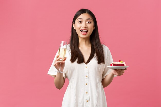 Celebrazione, feste di festa e concetto di divertimento. Bella donna sognante che festeggia il suo compleanno con un bicchiere di champagne e una torta di b-day, dall'aspetto sorpreso ed eccitato, in piedi su uno sfondo rosa.