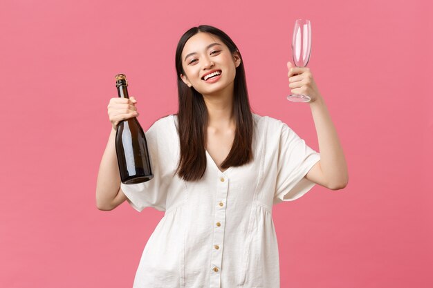 Celebrazione, feste di festa e concetto di divertimento. Gioiosa ragazza asiatica felice pronta a godersi il giorno libero con le amiche, portare champagne e bicchieri, macchina fotografica sorridente, in piedi sfondo rosa ottimista.