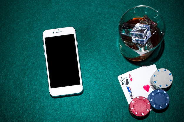 Cellulare e bicchiere di whisky con jack di vanga e carte asso cuore sul tavolo da poker verde