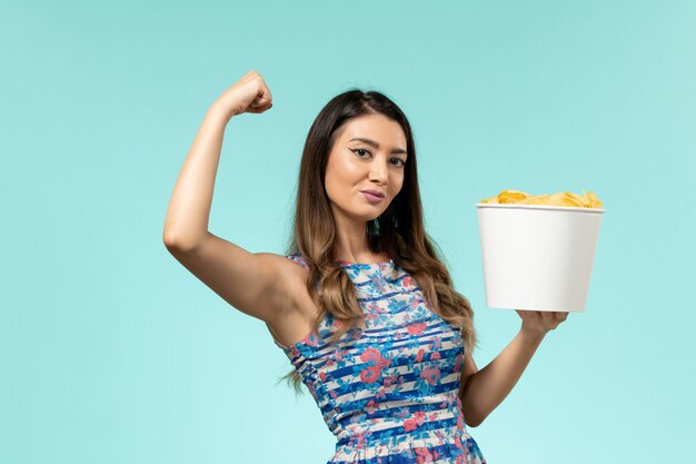 Cestino della holding della giovane femmina di vista frontale con le patatine fritte e che flette sulla superficie blu