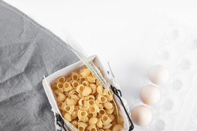 Cesto di pasta cruda e uova in contenitore sulla superficie bianca.