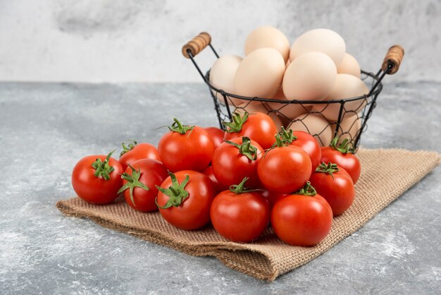 Cesto di uova crude fresche e pomodori maturi su marmo.