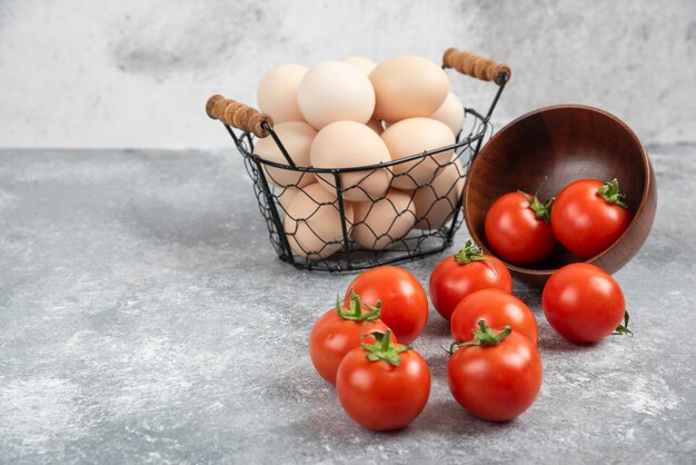 Cesto di vimini di uova biologiche crude e ciotola di pomodori su marmo.