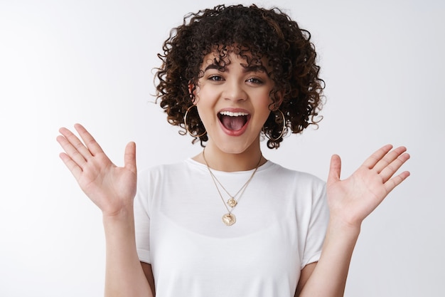 Che bella sorpresa. Stupito felice amichevole affascinante dai capelli ricci femmina bruna invitante collega felice di vedere amico sorridente alzando le mani stupito in piedi divertito felice intrattenuto, sfondo bianco