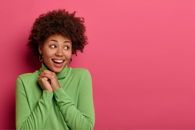 Che carino! La bella donna positiva ha folti capelli afro, si allontana, ammira qualcosa di molto piacevole, tiene le mani unite, sorride ampiamente, ha la pelle scura e sana