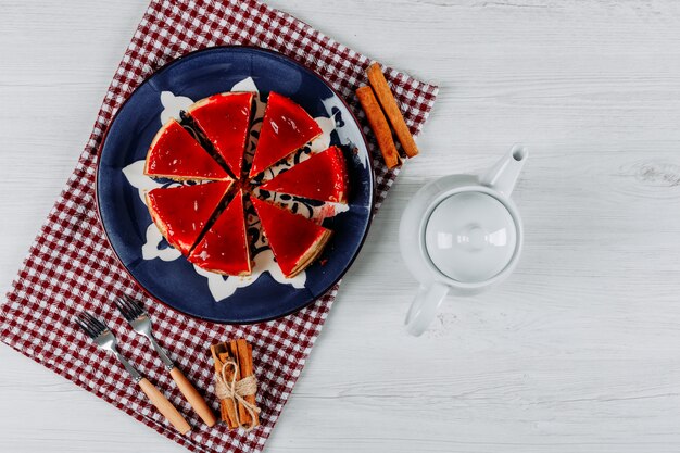 Cheesecake a fette in un piatto e un panno da picnic con una teiera, due forchette e bastoncini di cannella distesi su uno sfondo grigio chiaro